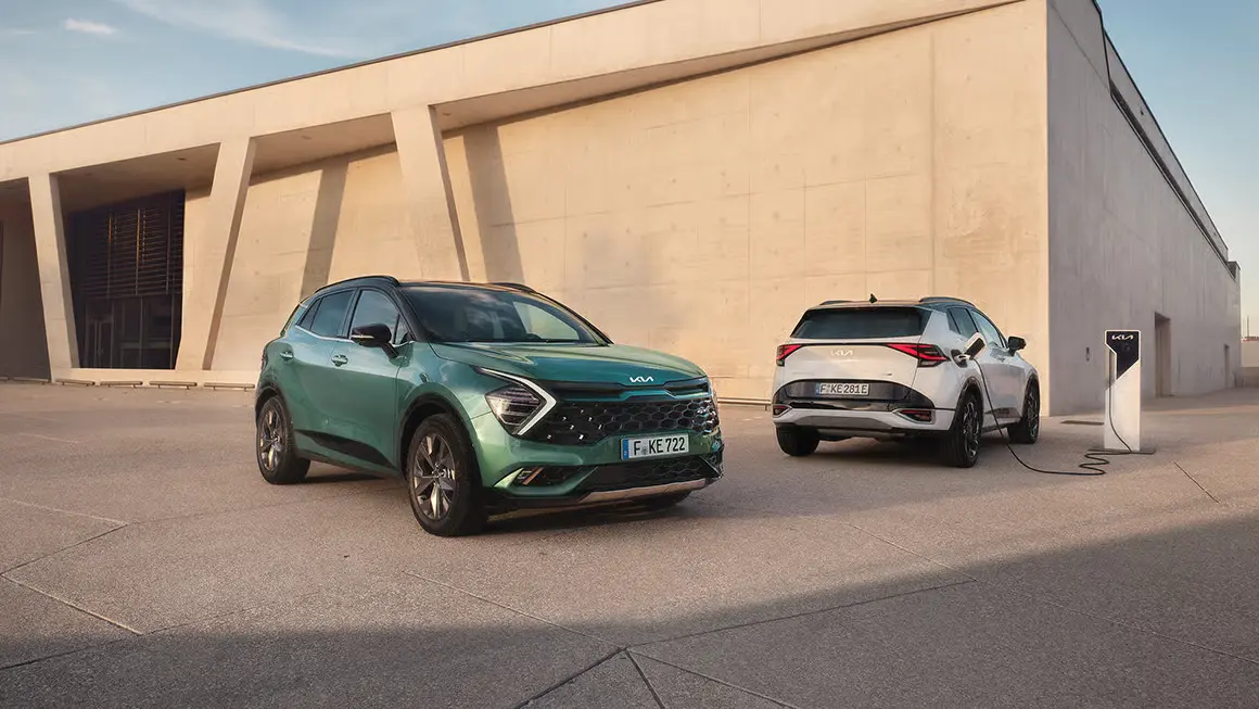 Zwei Kia Sportage Autos stehen vor einem modernen Gebäude, eines lädt an einer Ladestation.
