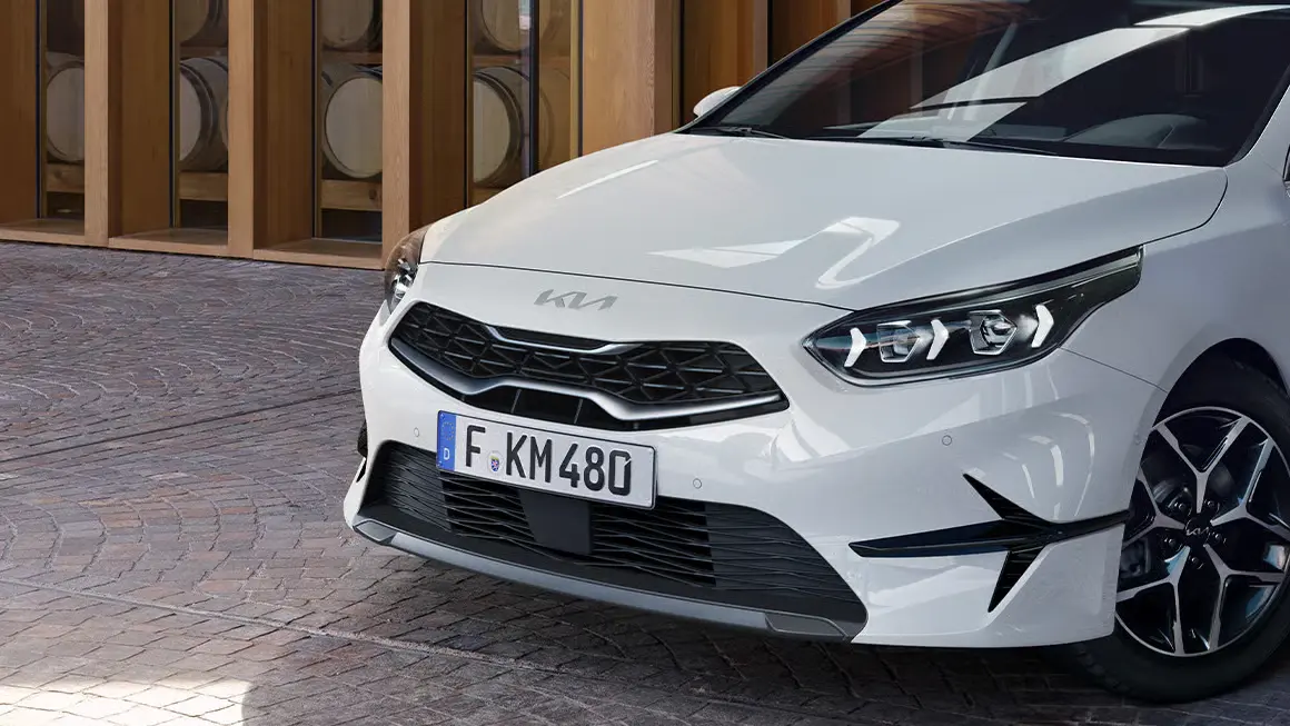 Ein weißer Kia Ceed SW steht auf gepflastertem Boden vor einem Gebäude.