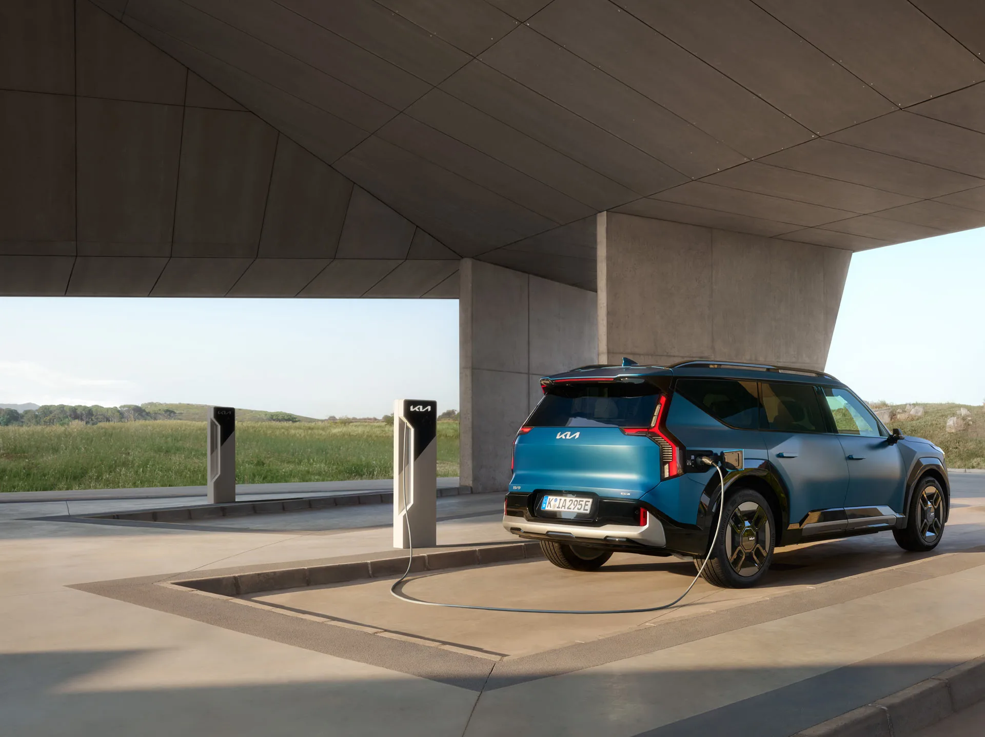Ein blauer Kia EV9 lädt an einer Ladestation unter einem modernen Betondach.