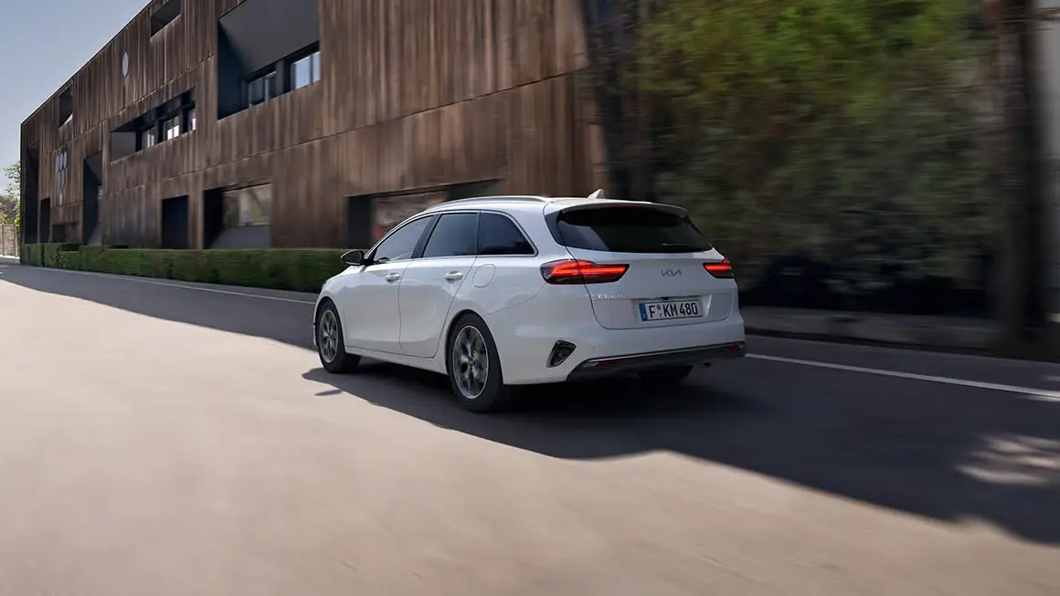 Ein weißer Kia Ceed SW fährt auf einer Straße entlang eines modernen Gebäudes.