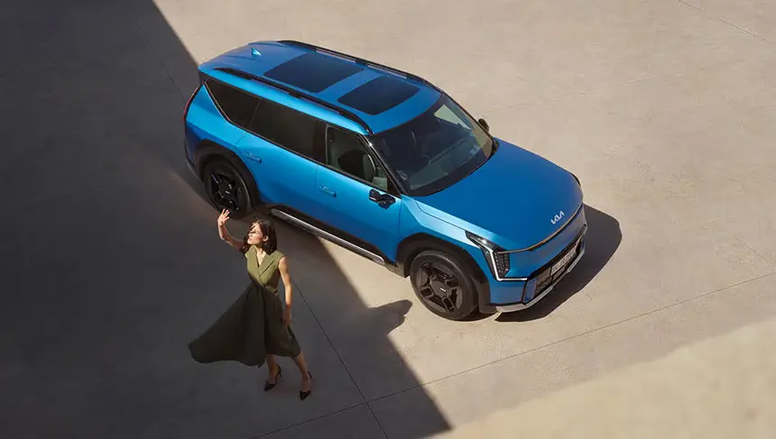 Eine Frau geht neben einem blauen Kia EV9 auf einem sonnigen Platz.