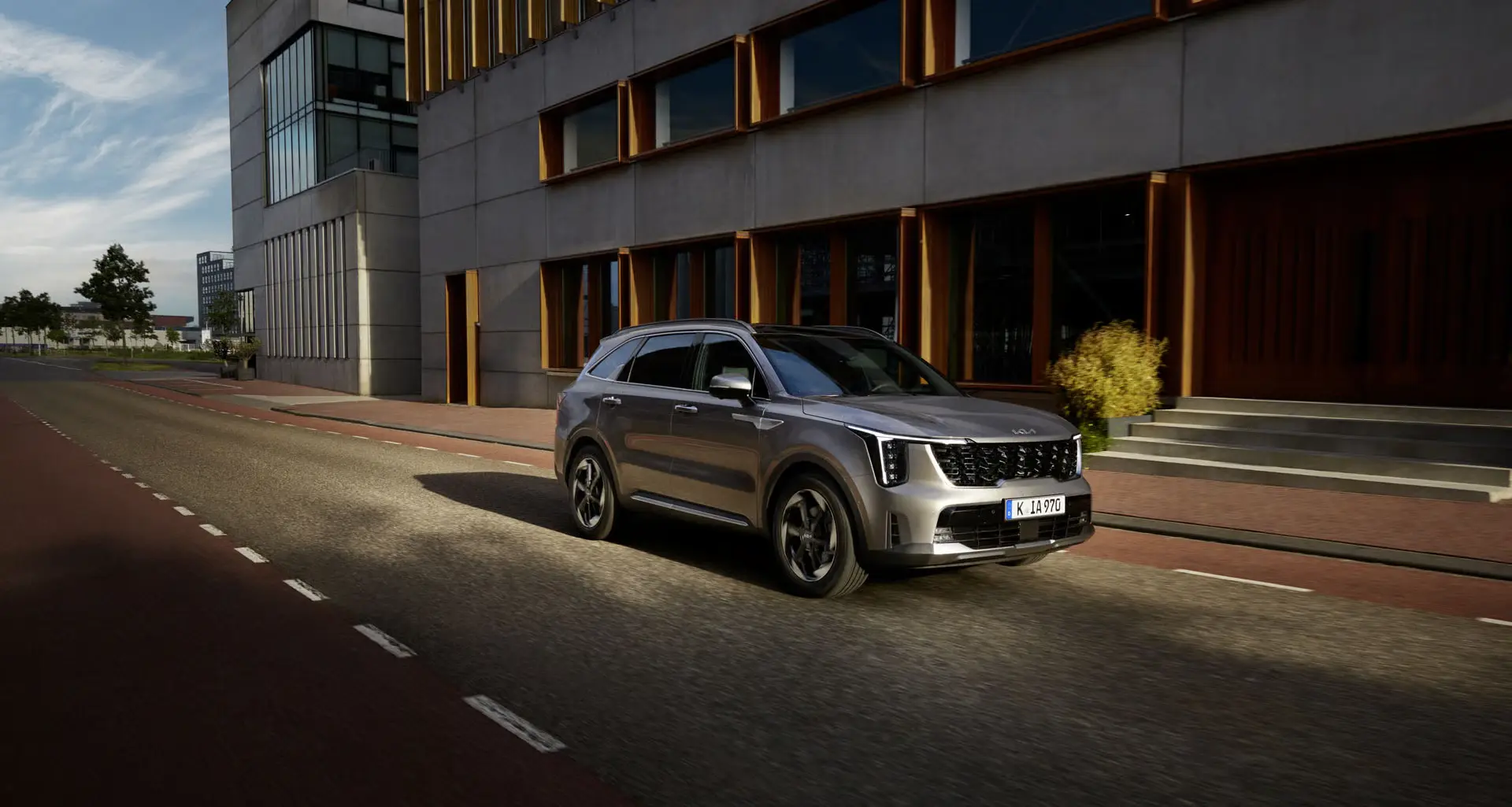 Ein silberner Kia Sorento fährt auf einer städtischen Straße entlang eines modernen Gebäudes.