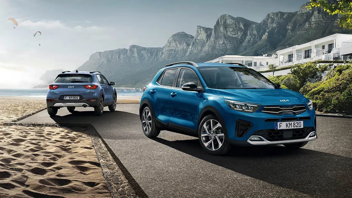 3-Slider-kia-stonic Zwei blaue Kia Stonic SUVs fahren auf einer Küstenstraße mit Bergen im Hintergrund.