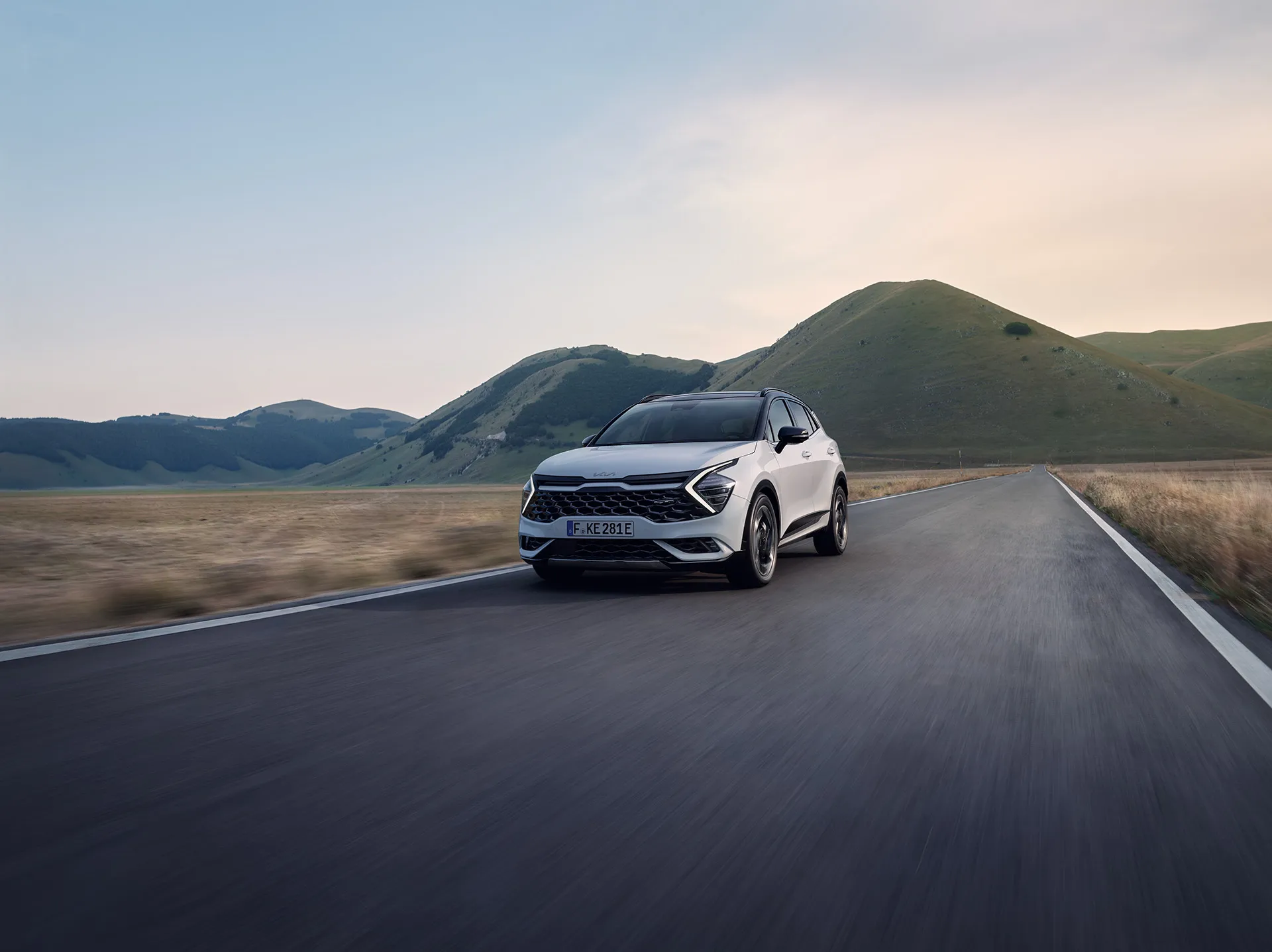 Ein weißer Kia Sportage fährt auf einer Landstraße vor einer hügeligen Landschaft.