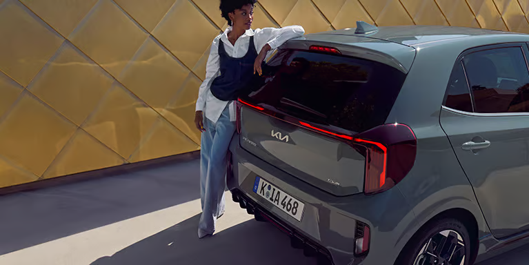 Eine Person lehnt an einem grauen Kia Picanto vor einer goldenen Wand.