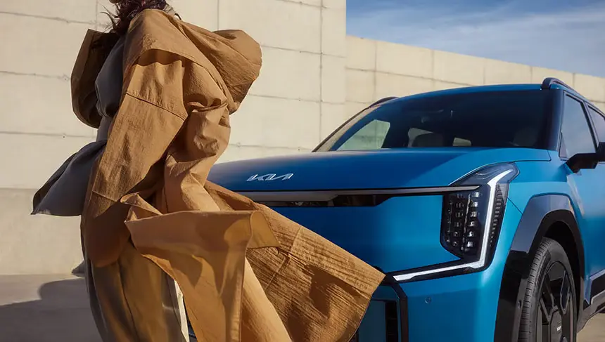 Eine Person in einem braunen Mantel steht neben einem blauen Kia EV9.