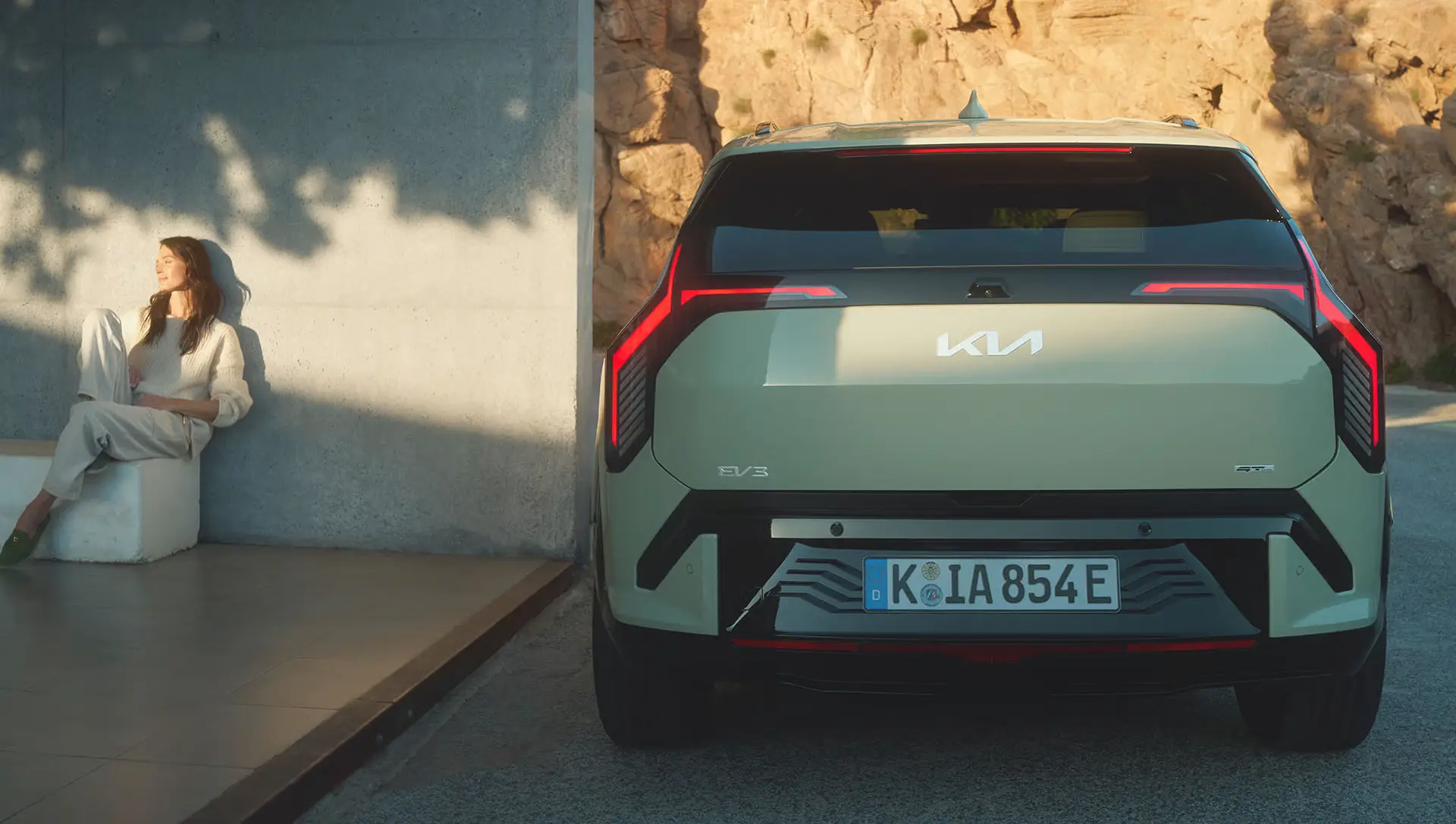 Rückansicht eines Kia EV3 neben einer entspannt sitzenden Person an einer Wand.