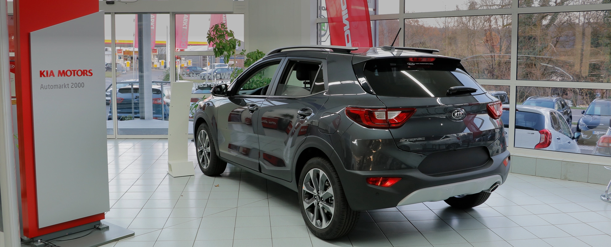 Ein grauer Kia SUV steht in einem Autohaus neben einem Schild mit der Aufschrift "KIA MOTORS Automarkt 2000".