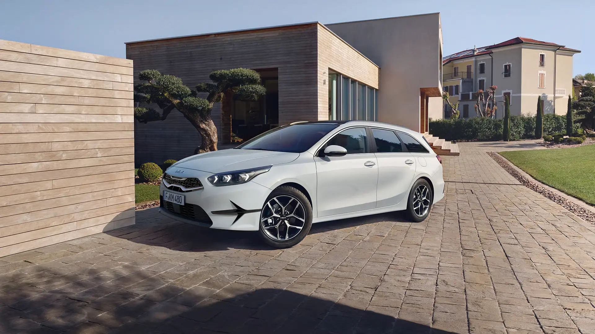 Weißer Kia Ceed SW auf einer gepflasterten Auffahrt vor einem modernen Haus.