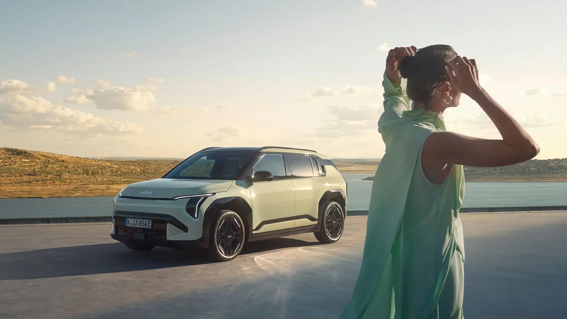 Eine Frau steht vor einem Kia EV3 SUV in einer landschaftlichen Umgebung.
