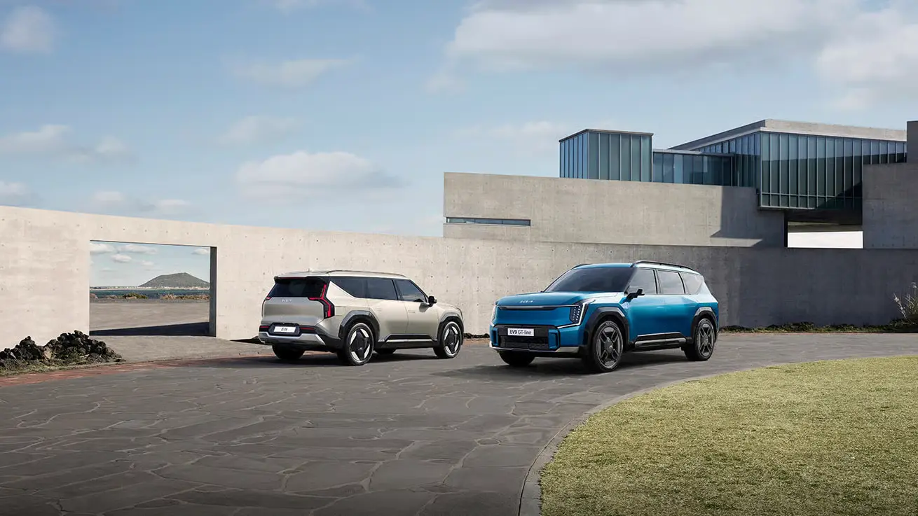 Zwei Kia EV9 SUVs, einer in Blau und einer in Beige, parken vor einem modernen Gebäude.
