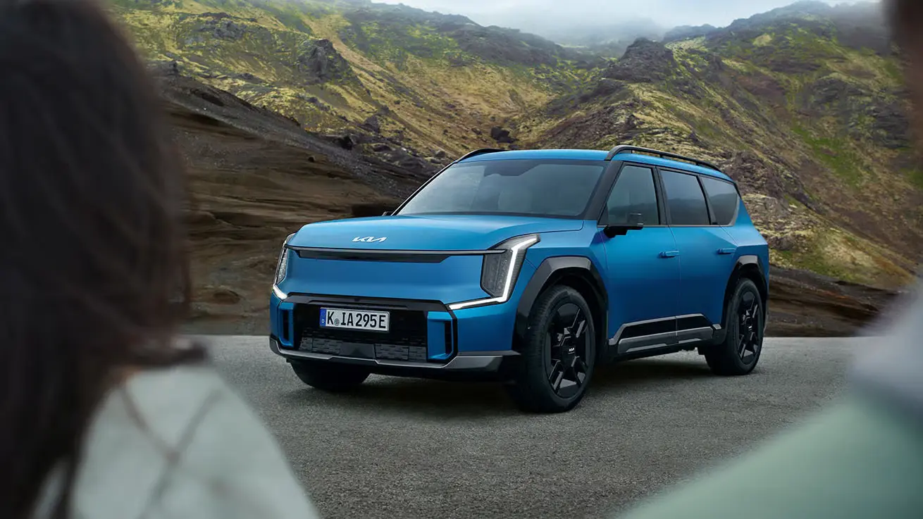 Blauer Kia EV9 SUV auf einer Straße vor einer bergigen Landschaft.
