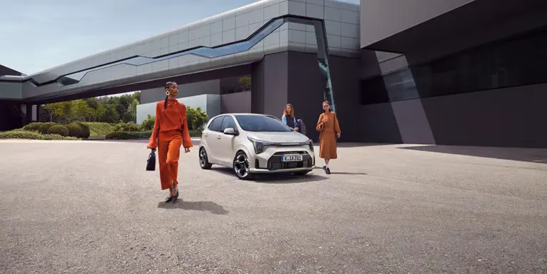 Ein weißer Kia Picanto steht vor einem modernen Gebäude, umgeben von drei Personen.