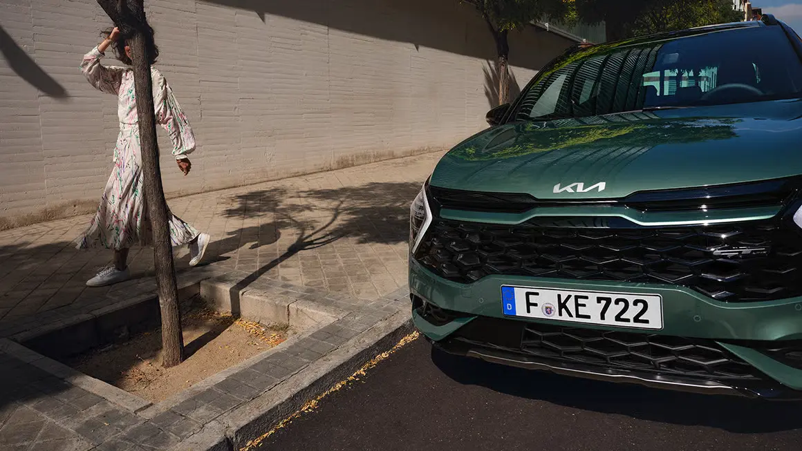 Ein grüner Kia Sportage HEV parkt am Straßenrand neben einer Person, die an einem Baum vorbeigeht.