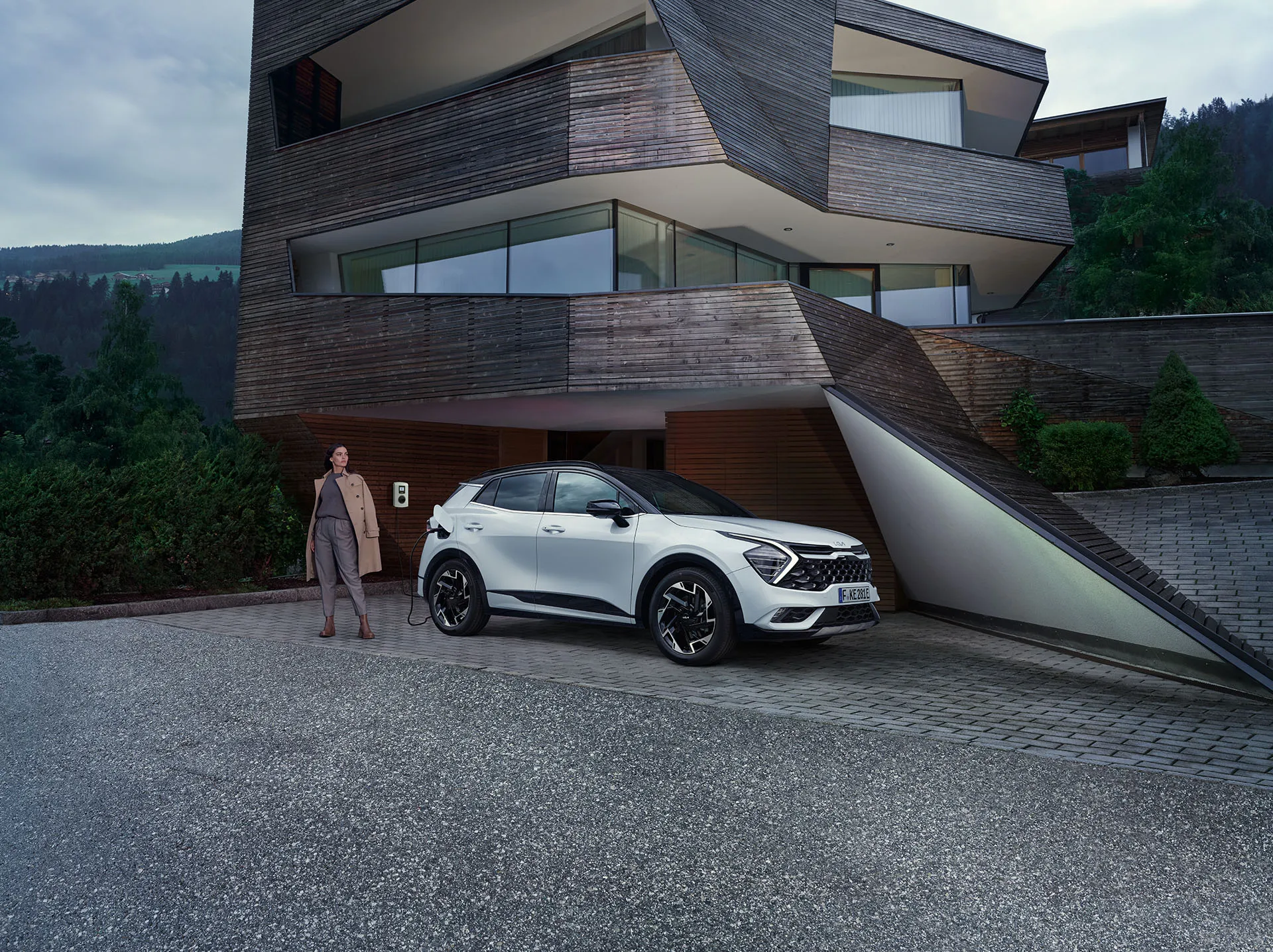 Eine Frau steht neben einem weißen Kia Sportage PHEV, der an einer Ladestation angeschlossen ist, vor einem modernen Gebäude.