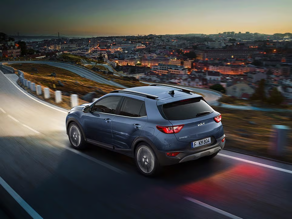 kia-stonic-my24-ecodynamics Ein blauer Kia Stonic fährt bei Dämmerung eine kurvige Straße entlang.