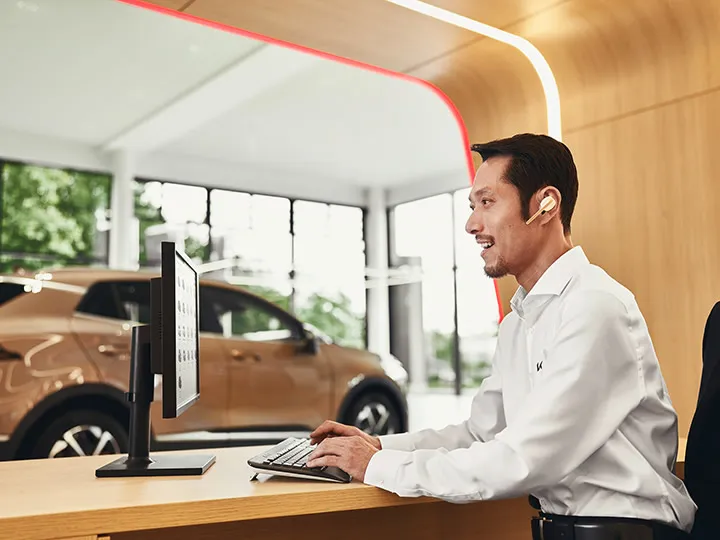 Ein Mann in einem Autohaus spricht am Computer, im Hintergrund steht ein KIA-Fahrzeug.