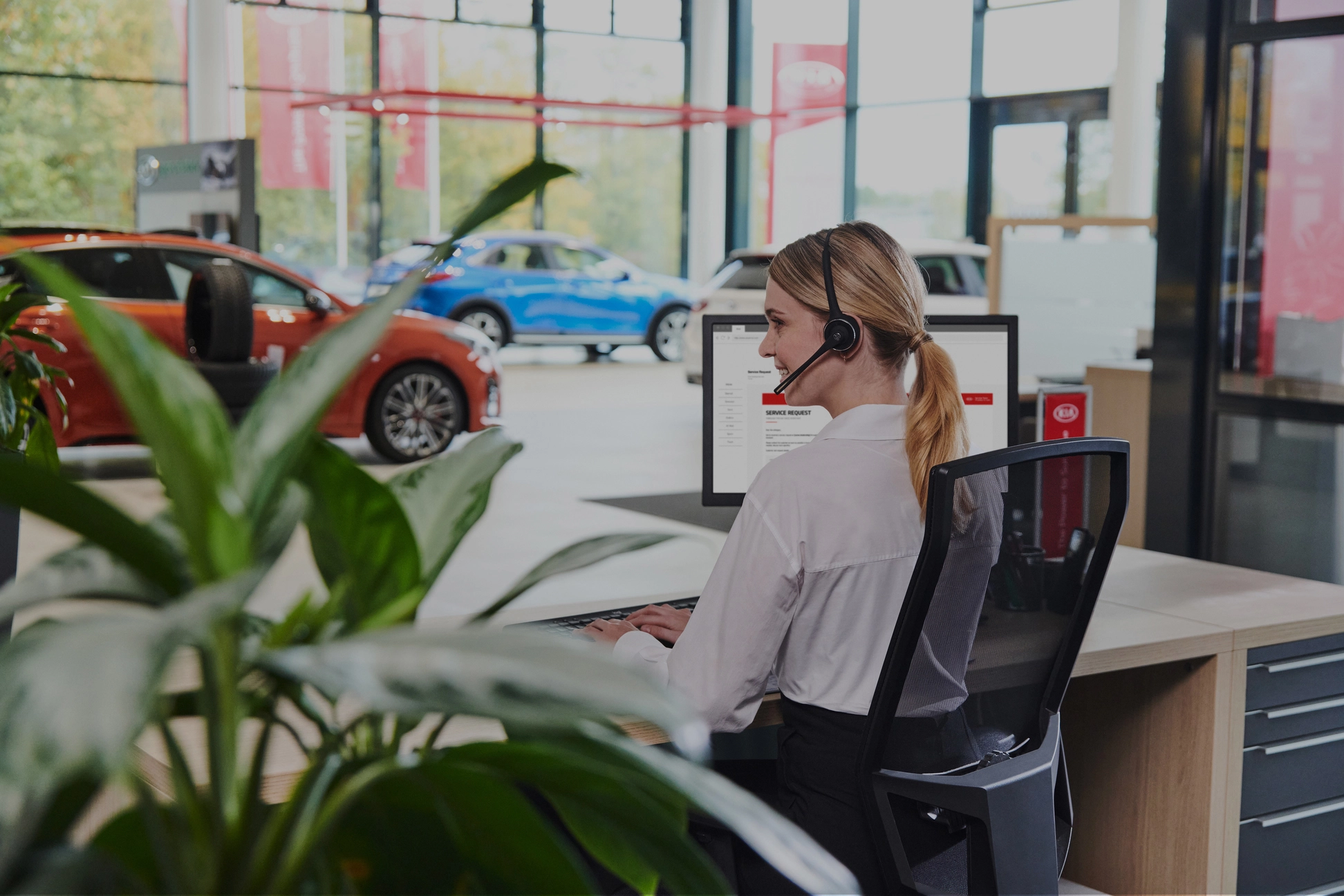 Eine Frau mit Headset sitzt an einem Schreibtisch in einem Autohaus, im Hintergrund sind Autos zu sehen.