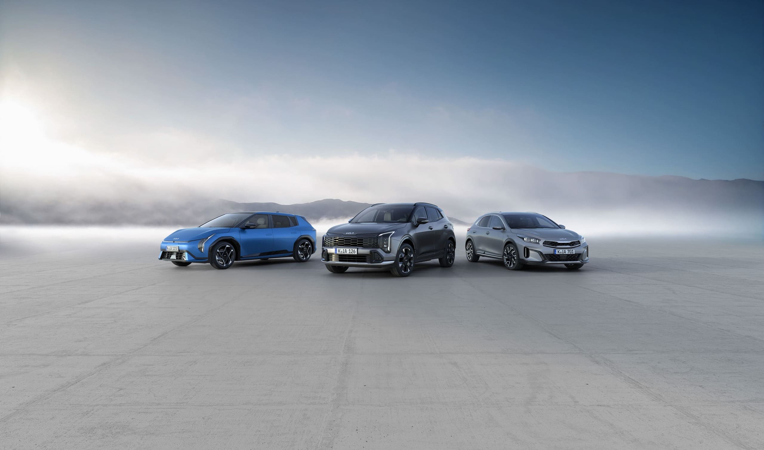 Drei Kia-Fahrzeuge (SUVs/Limousine) in Blau, Grau und Silber vor nebliger Fläche.